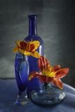 Blue Glass and Bottles with two orange and red flowers against a blue grey background..